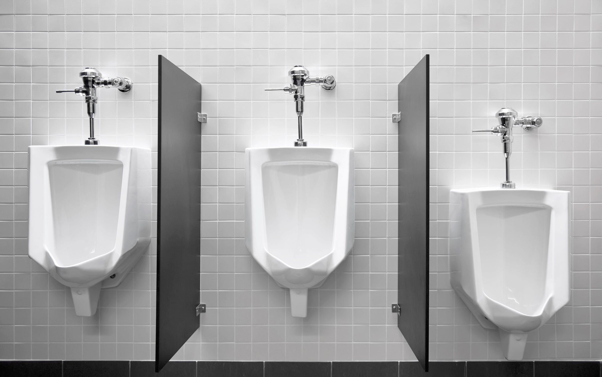 Row of three wall-hung urinals with flush valves and privacy dividers on a white tile wall